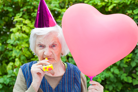 Picture of a happy elderly woman celebrating birthday holding a heart shaped balloon an blowing a whistle outdoorの写真素材