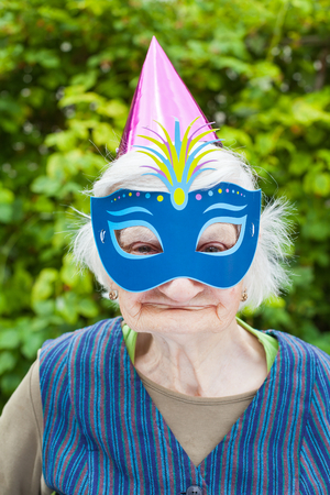 Portrait of a happy elderly woman celebrating birthday outdoor, wearing a colorful hat and maskの写真素材