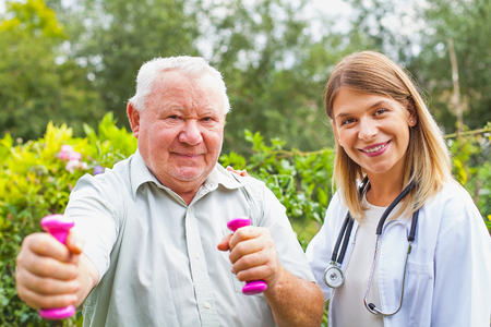 Smiling female therapist doing strength exercises with dumbbell with elderly male patient in the...