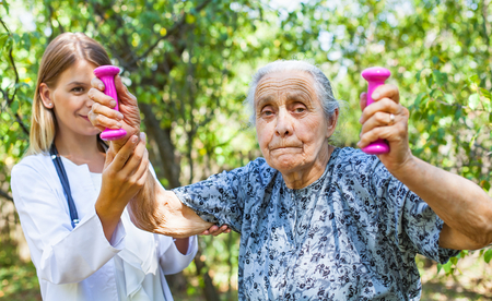 Elderly female doing exercises with dumbbells in the park assisted by young female physicianの写真素材
