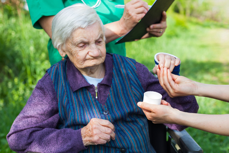 Elderly ill woman receiving pills from nurse outdoor, doctor writing medical prescription in the backgroundの写真素材