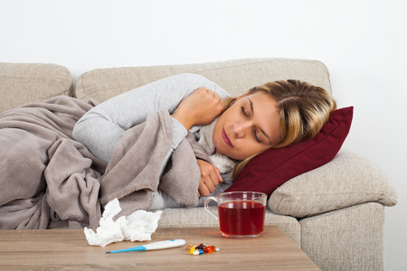 Sick woman lying on the couch wrapped in a warm blanket - medication, thermometer and tea on the tableの写真素材