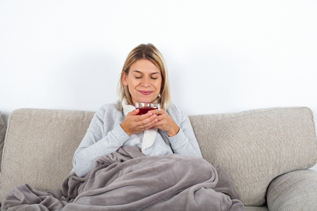 Blonde attractive woman drinking hot tea while sitting on the couch, wrapped in a grey blanketの写真素材