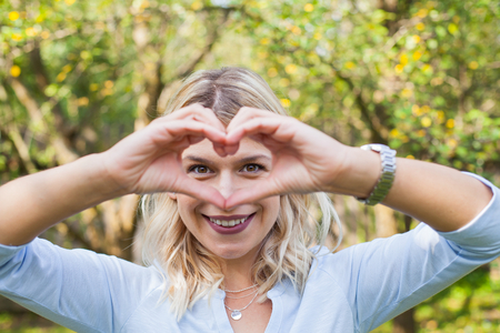 Charming young lady showing a heart symbol with her hands, looking to the camera outdoorの写真素材