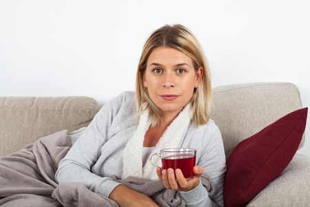 Beautiful woman relaxing on the sofa and drinking hot tea, wrapped in a grey blanketの写真素材