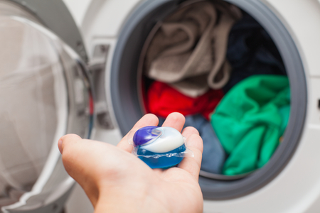 Close up picture of person holding laundry detergent 3 in 1 pod capsule, washing machine with laundry in the backgroundの写真素材