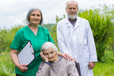 Aged woman with dementia sitting in a wheelchair, male doctor and female nurse taking care of her outdoor - assisted livingの写真素材