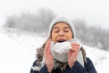 Portrait of a young woman sneezing , holding a tissue outdoor in wintertimeの写真素材