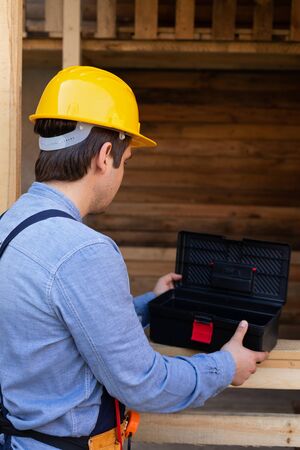 Back view of young carpenter with yellow hard hat and blue shirt outdoor, wooden background
