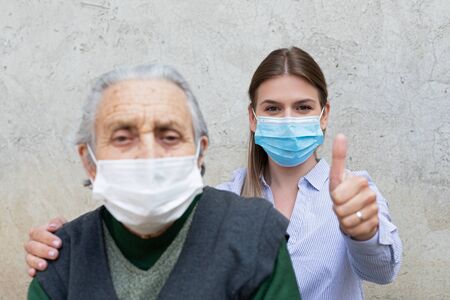 Portrait of friendly caregiver posing with elderly ill woman wearing surgical mask because of covid-19 pandemic, showing thumbs upの素材 [FY310144858880]