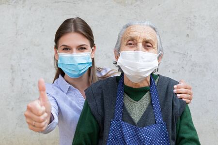 Portrait of friendly caregiver posing with elderly ill woman wearing surgical mask because of covid-19 pandemic, showing thumbs upの写真素材
