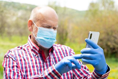 Portrait of elderly man wearing surgical mask and gloves talking on the phone outdoorsの写真素材
