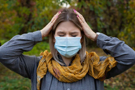 Young caucasian woman with surgical mask having headache and fever outdoor. Covid-19 symptoms