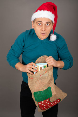 Young handsome man with Santa hat is holding a bag full of Christmas gifts in front of grey...