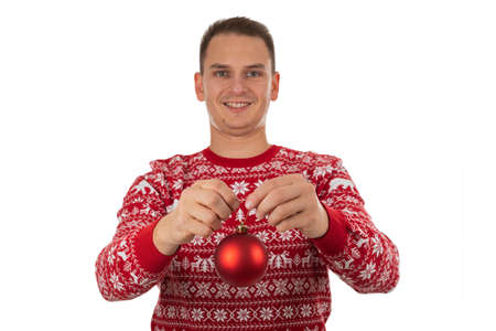 Young cheerful man wearing winter style knitwear is holding a red Christmas globe on isolated backgroundの写真素材