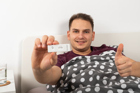 Smiling young man in bed at home with covid-19 symptoms is holding a quick coronavirus test and...