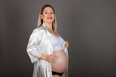 Beautiful young mom to be posing in front of grey background - Pregnancy photography wearing elegant underwear indoorの写真素材