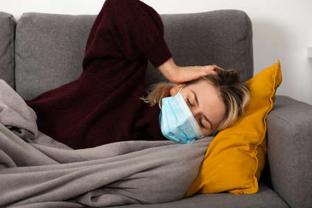 Young woman with coronavirus is having headache and fever, lying on the sofa wrapped in a balnketの写真素材