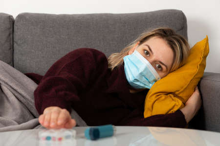 Young woman with coronavirus is having headache and fever, lying on the sofa wrapped in a balnket