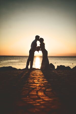 Silhouette of a loving, married couple where he is kissing her hand. In the background of the setting sun, rocks and sea.の写真素材