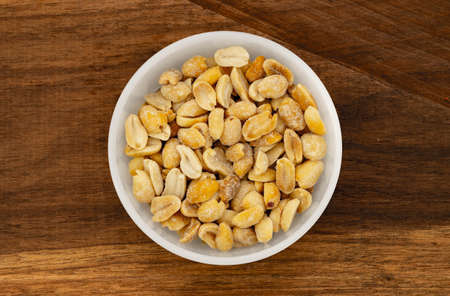 Salted peanuts in white bowl, photographed overhead on dark wood with natural light. Top view of...