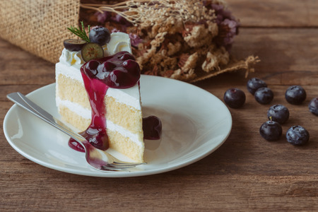 Blueberry cream cake on white plate on wood table in side view. Vanilla sponge cake decorated with dairy whipped cream and blueberry sauce so soft sweet and delicious. Homemade bakery concept.の写真素材