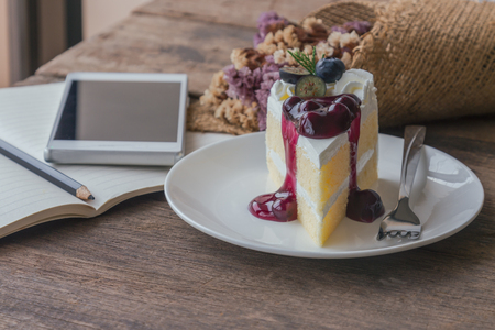 Blueberry cream cake on wood table beside window near notebook and smart phone . Vanilla sponge cake with dairy whipped cream and blueberry sauce so soft sweet and delicious. Homemade bakery concept.の写真素材
