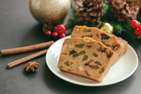 Sweet fruit cake slices on white plate put on black granite table in side view with Christmas decoration. Traditional rum fruit cake for Christmas celebrate festival. Homemade bakery concept.の写真素材