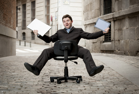 Happy crazy business man rolling downhill on chair with computer and tabletの写真素材