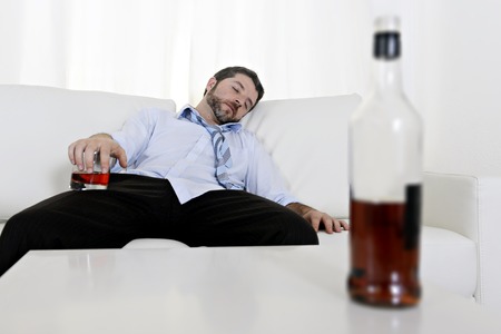  drunk business man at home lying asleep on couch sleeping wasted holding whiskey glass in alcoholism problem , alcohol abuse and addiction concept looking messy and sickの写真素材