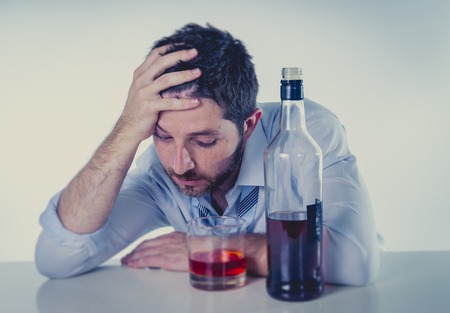 young alcoholic business man with beard looking wasted suffering hangover wearing blue shirt and tie drunk and drinking   Whisky on office desk at work cool color editの写真素材