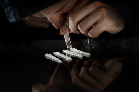 young drug addict man on hood alone sniffing and snorting cocaine lines with rolled banknote on mirror in moody and grunge lighting studio settingの写真素材