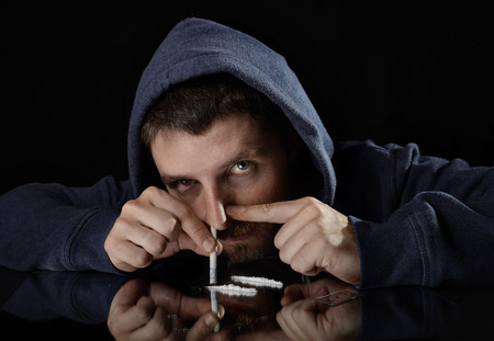 young drug addict man on hood alone sniffing and snorting cocaine lines with rolled banknote on mirror looking with sick red eyes in moody and grunge lighting studio settingの写真素材