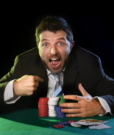 happy young attractive man grabbing poker chips pointing himself with his finger after winning bet gambling on table with playing cards on green felt at Casinoの写真素材