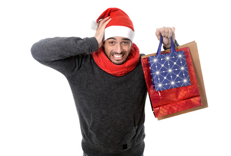young attractive shopaholic man wearing santa hat holding a lot of shopping bags looking desperate and in stress in christmas consumerism, sale, compulsive buying, spending money and xmas gifts and presents concept isolated on white backgroundの写真素材