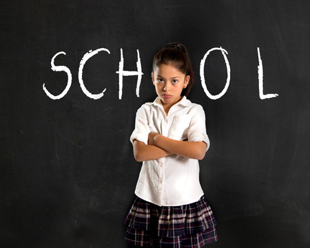 latin moody little girl in uniform standing upset in front of blackboard with school written with...
