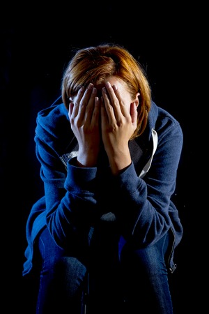 young woman suffering depression and stress sitting alone in pain and grief covering face with hands feeling sad and desperate isolated on black backgroundの写真素材
