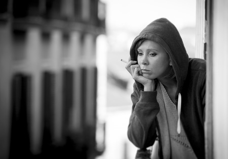 young attractive woman suffering depression and smoking in stress outdoors at home balcony terrace window in pain and grief feeling sad and desperate in urban background in black and whiteの写真素材