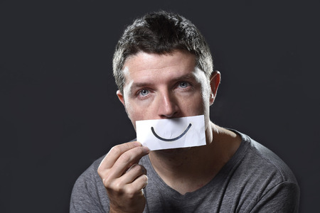 young depressed man lost in sadness and sorrow holding paper with smiley on his mouth as society...