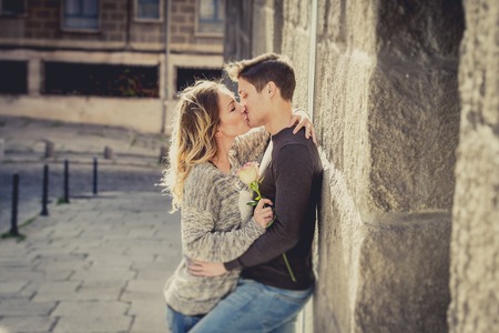 candid portrait of beautiful European couple with rose in love kissing on street alley celebrating Valentines day with passion against stone wall on urban backgroundの写真素材