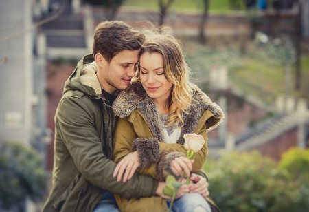 candid portrait of beautiful European couple with rose in love kissing on street  celebrating Valentines day with passion sitting on city park in winter urban backgroundの写真素材