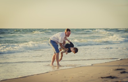 Young happy father holding in his arms little son playing on the beach with barefoot in sand and water, the kid smiling and having fun together with dad in Summer vacation conceptの写真素材
