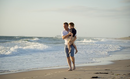 Young happy father holding in his arms little son walking on the beach with barefoot in sand in front of sea waves, the kid smiling and having fun together with dad in Summer vacation conceptの写真素材