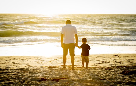 Young happy father holding holding hand of little son walking together on the beach with barefoot in sand in front of sea waves, the kid smiling and having fun  with dad in Summer sunset coastの写真素材