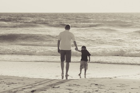 Young happy father holding holding hand of little son walking together on the beach with barefoot in sand in front of sea waves, the kid smiling and having fun  with dad in Summer sunset coastの写真素材