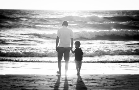 Young happy father holding holding hand of little son walking together on the beach with barefoot in sand in front of sea waves, the kid smiling and having fun  with dad in Summer sunset coastの写真素材