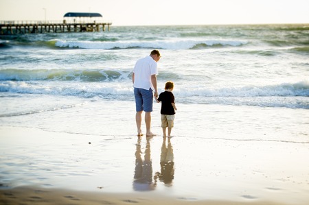 young happy father holding hand of little son walking together on the beach with barefoot in sand in front of sea waves, the kid smiling and having fun  with dad in Summer sunset coastの写真素材