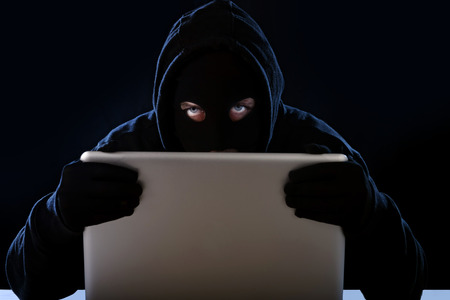 hacker man in black hood and mask with computer laptop in dangerous dark look hacking system having access to data info and privacy in business digital intruder and cyber crime conceptの写真素材