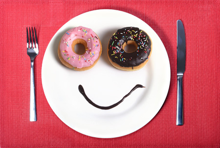 smiley happy face made on dish with donuts as eyes and chocolate syrup as smile in sugar and sweet addiction and nutrition concept on red mat with knife and forkの写真素材
