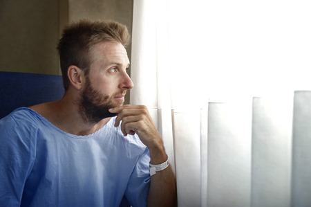 young injured man in hospital room sitting alone in pain looking on window negative and worried for his bad health condition sitting on chair suffering depression on sad lonely backgroundの写真素材